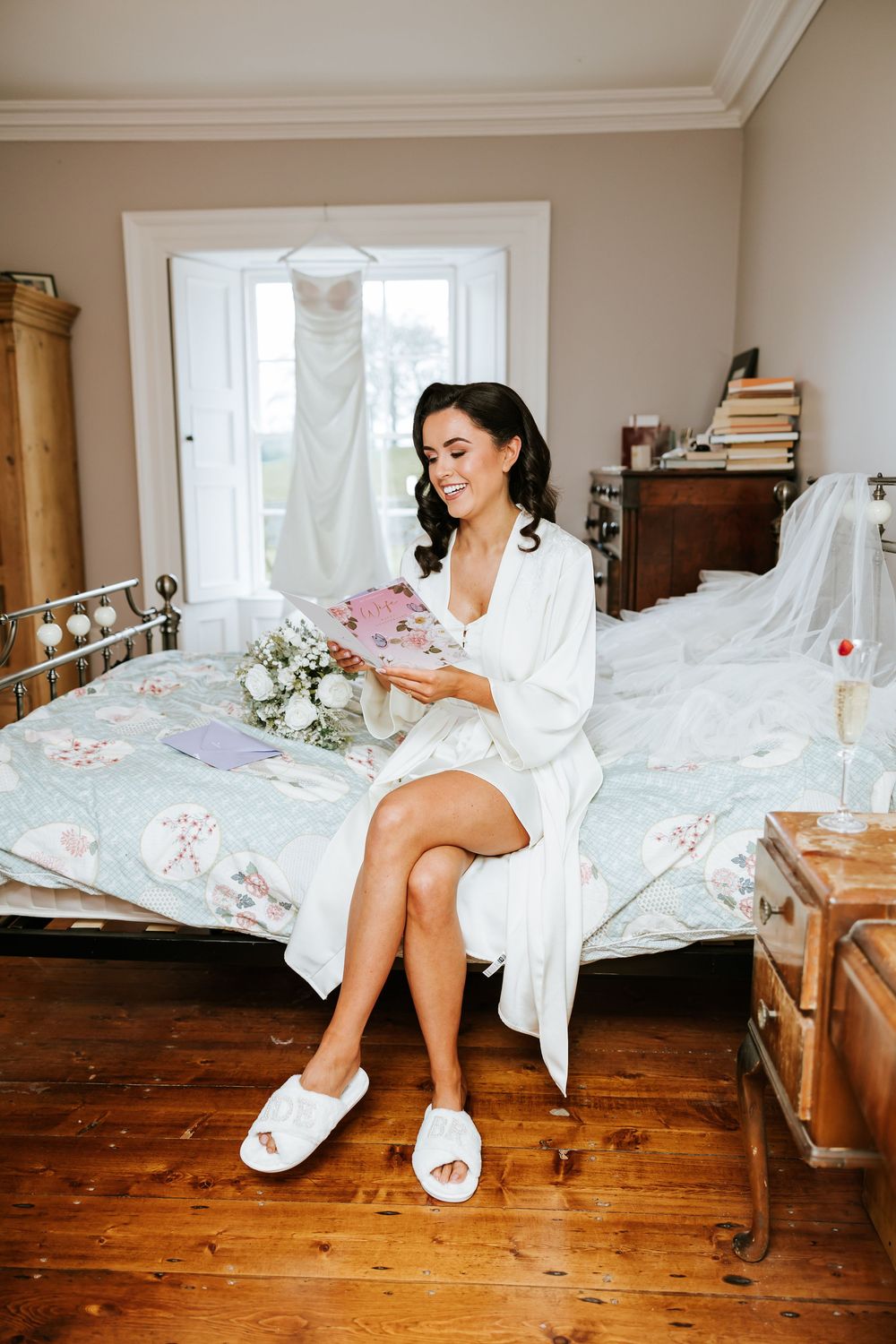Bride in white robe sits on wooden floor during wedding day preparations in sunlit room. Bellingham Castle Conlon Photo & Film