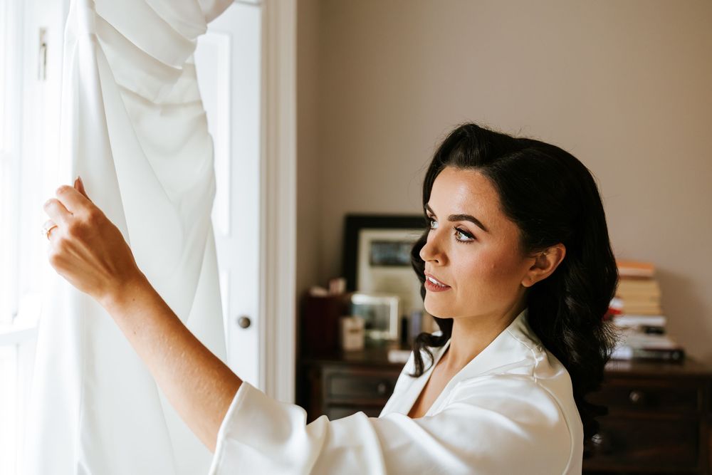 Woman in white shirt looking out window while adjusting curtains in sunlit room. Bellingham Castle Conlon Photo & Film