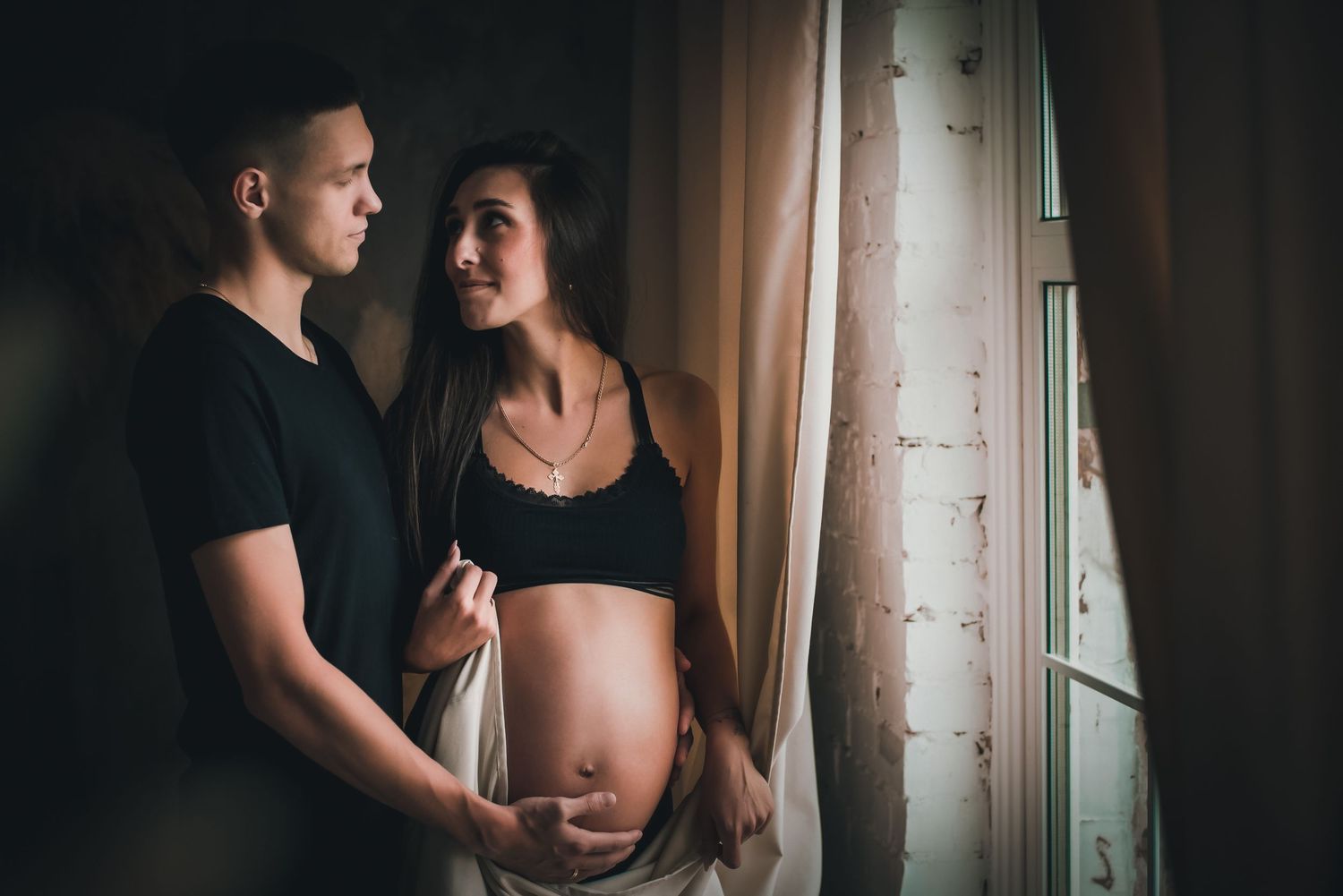 A couple in black attire shares an intimate moment during a maternity photoshoot near a window.
