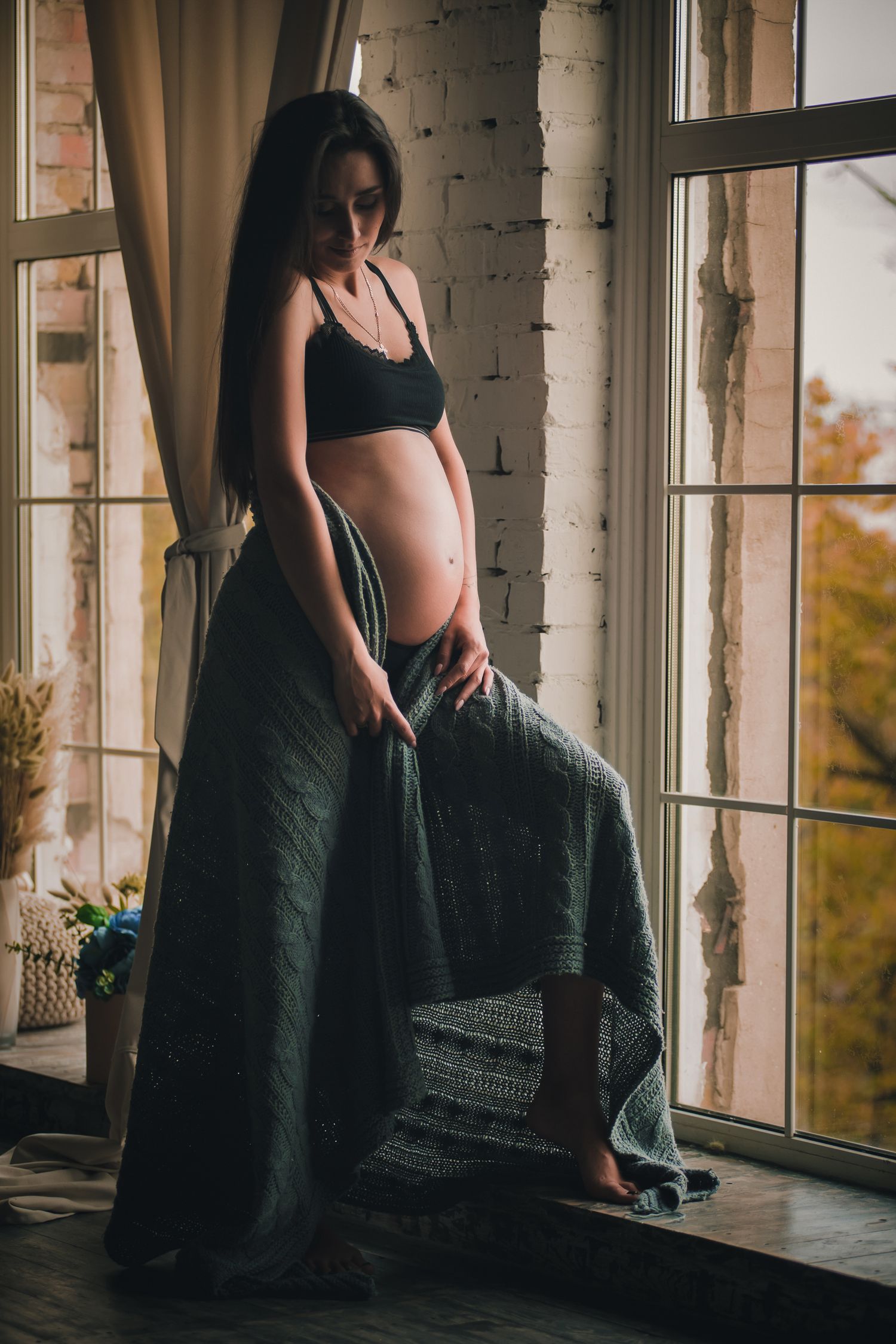 A silhouetted figure in black dress stands by large windows in a rustic room with brick walls and autumn views outside.