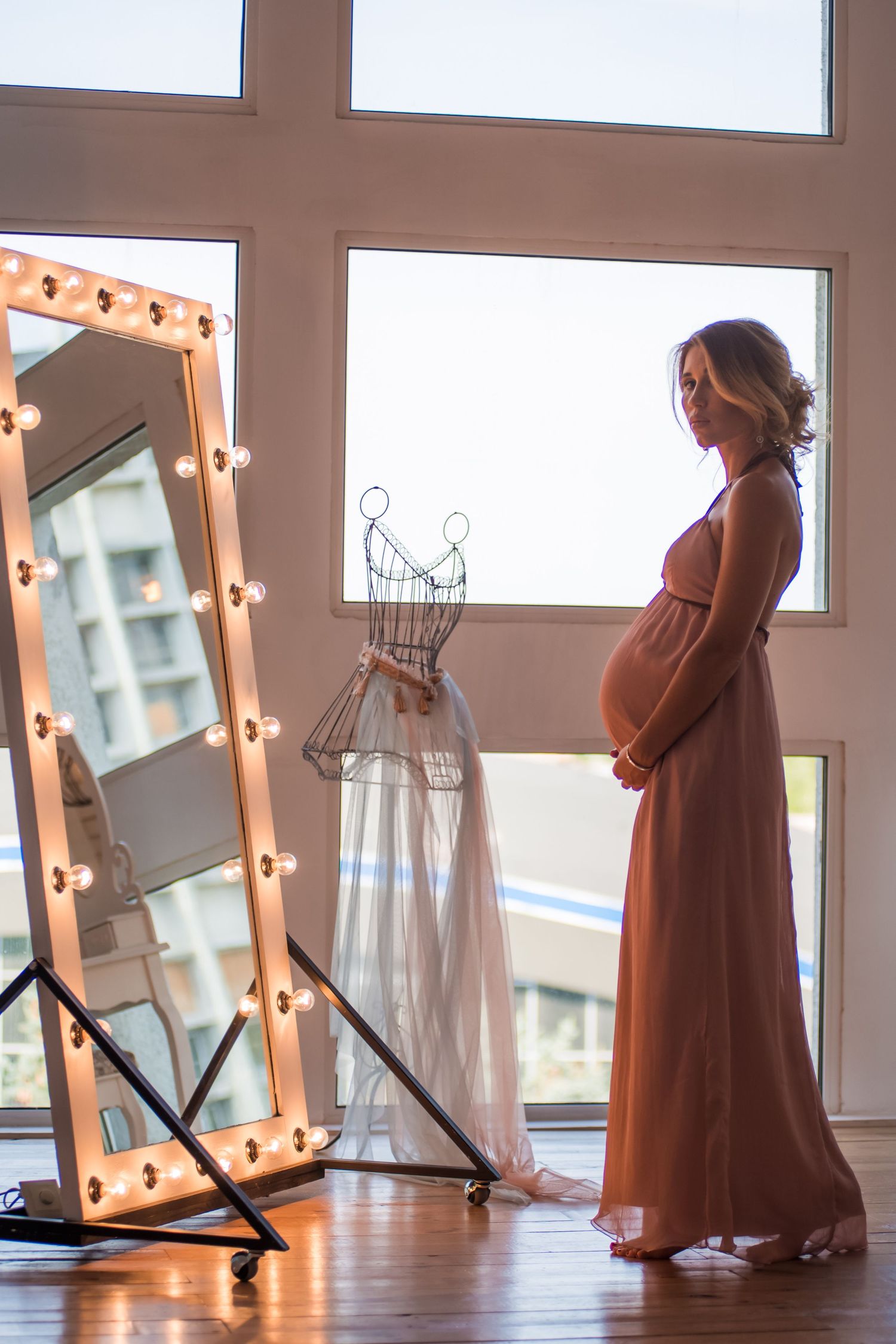 A series of photos showing a person in a rust-colored dress posing next to a lighted vanity mirror in a bright studio space.