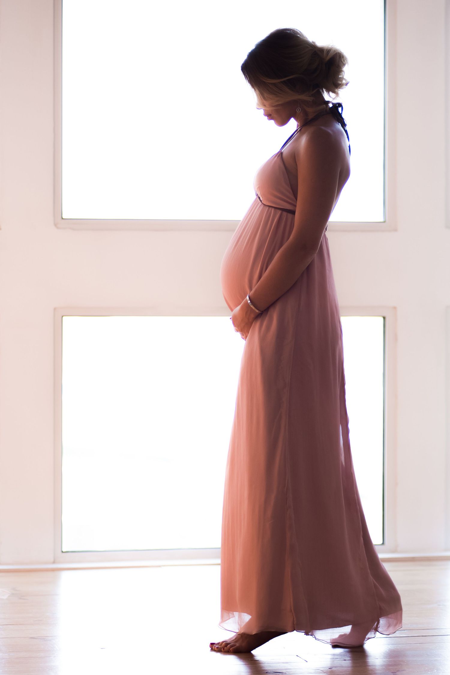 Silhouette of pregnant woman in flowing peach dress standing near bright windows in a minimalist white room.
