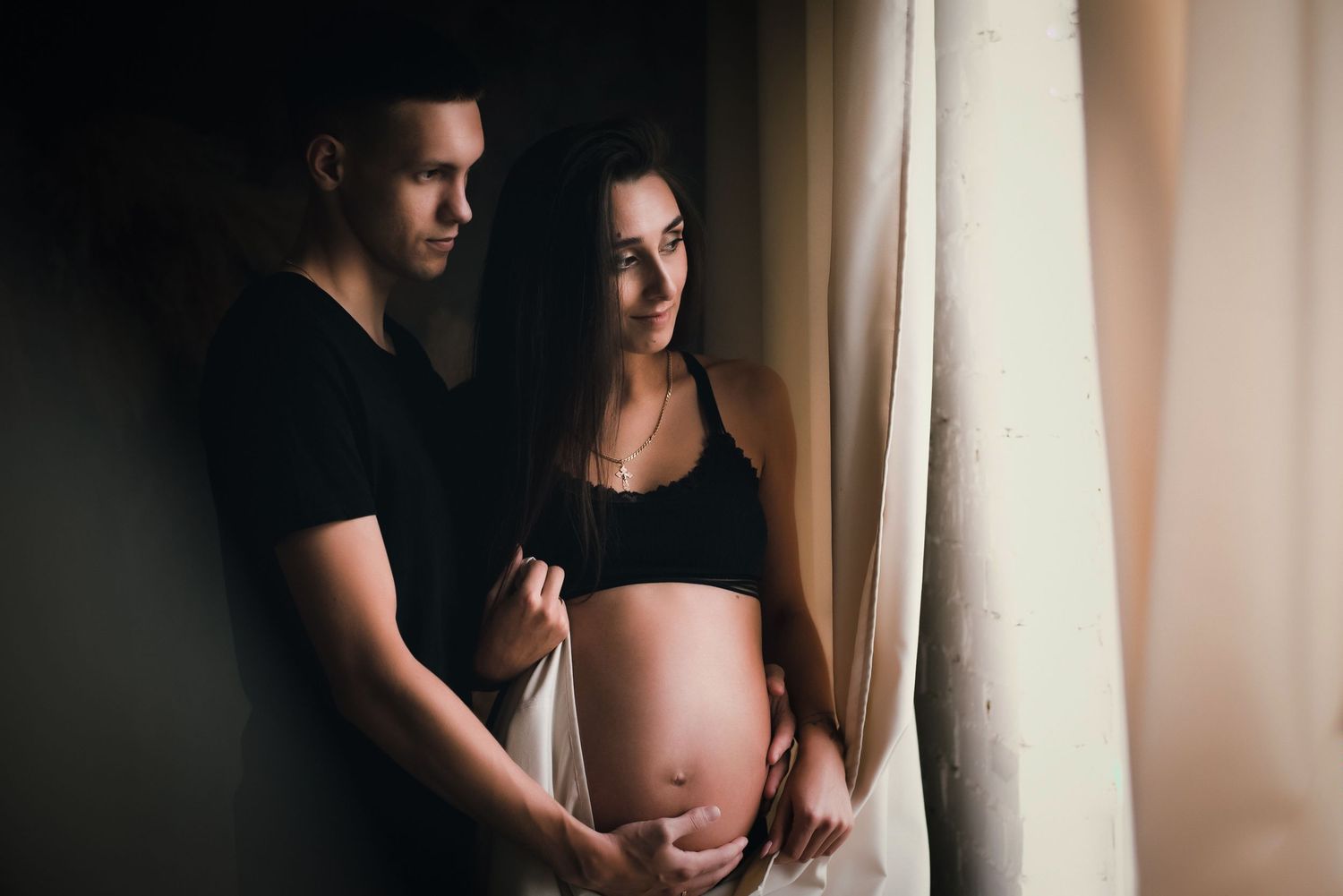 A couple stands by a window in an intimate maternity photo, with soft natural light illuminating the scene.