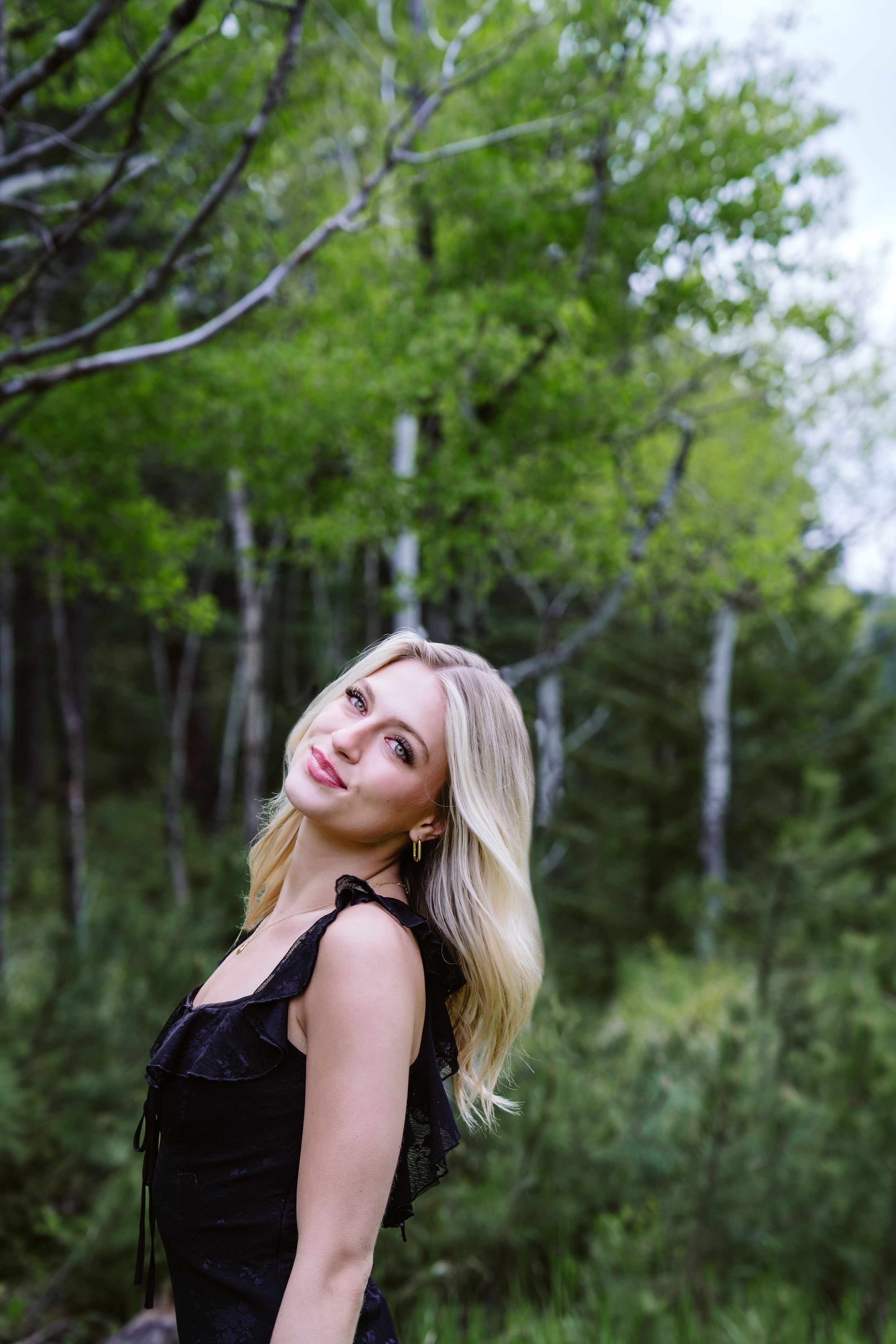 Person in black dress poses playfully against backdrop of green summer birch forest.