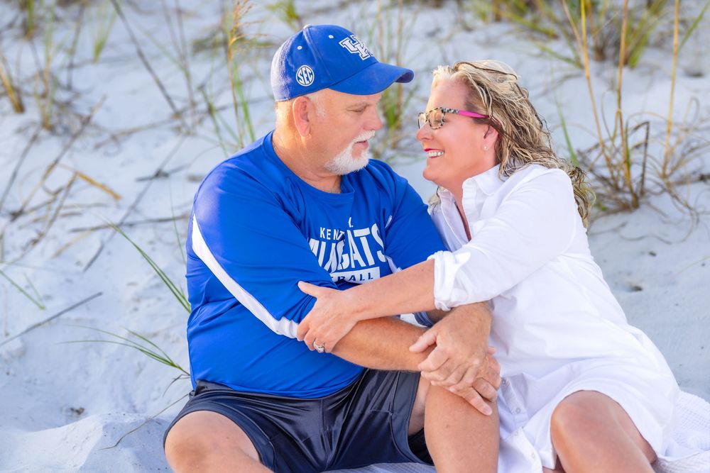 Couple in blue and white outfits share a tender moment together on a sandy beach during a sunny day.