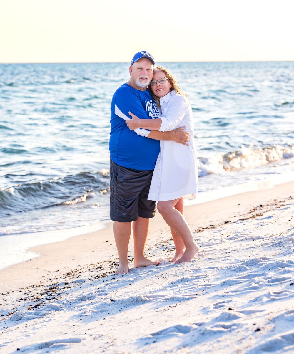 A couple embraces while standing on a sandy beach with ocean waves in the background on a sunny day.