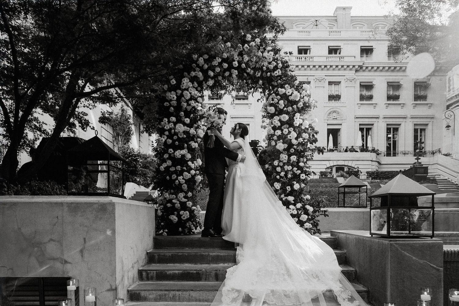 Black and white photo of a wedding couple under an elaborate floral arch at a grand estate.