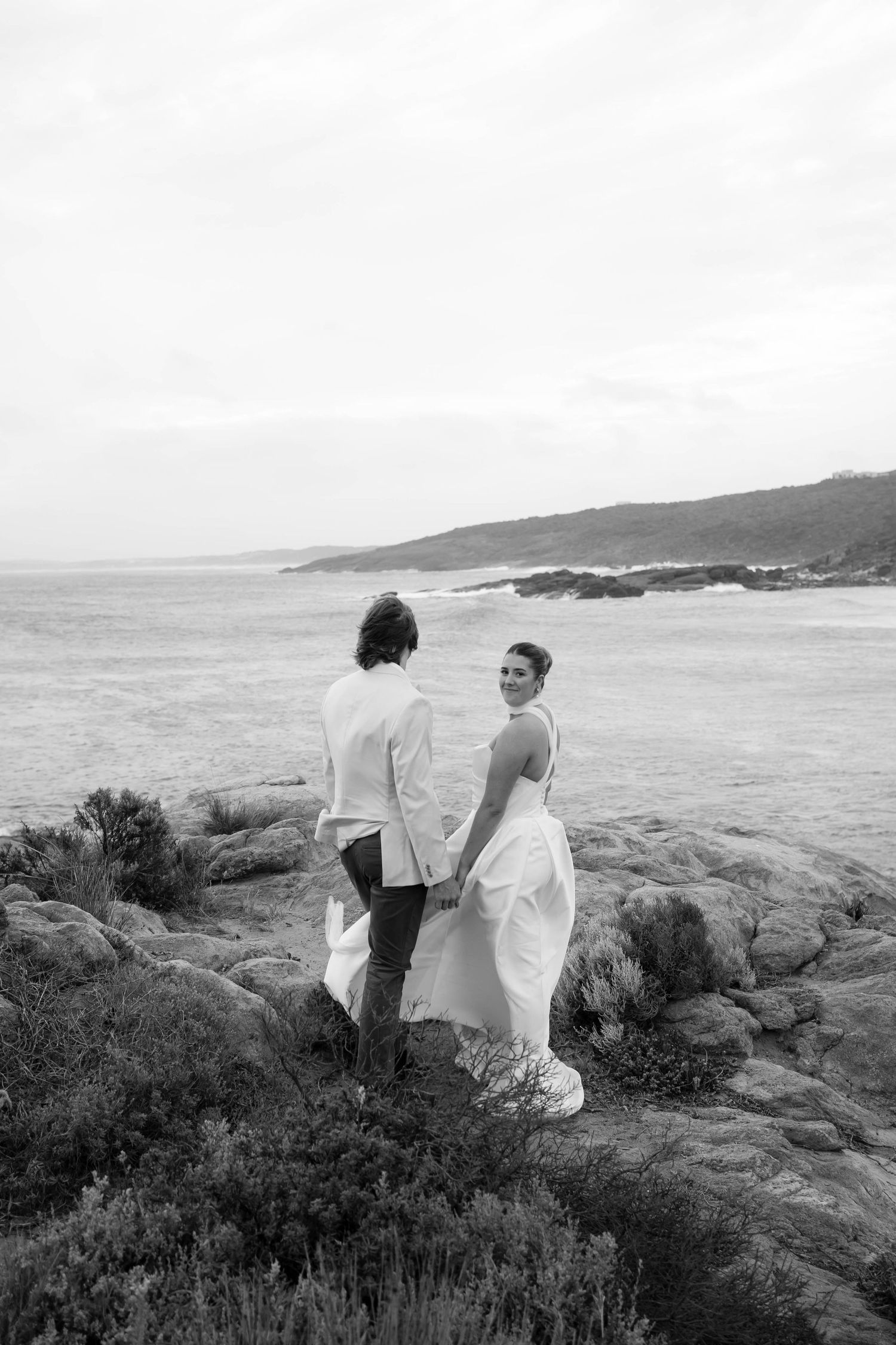 Two people in white dresses stand on rocky coastal cliffs overlooking the ocean in a black and white photograph.