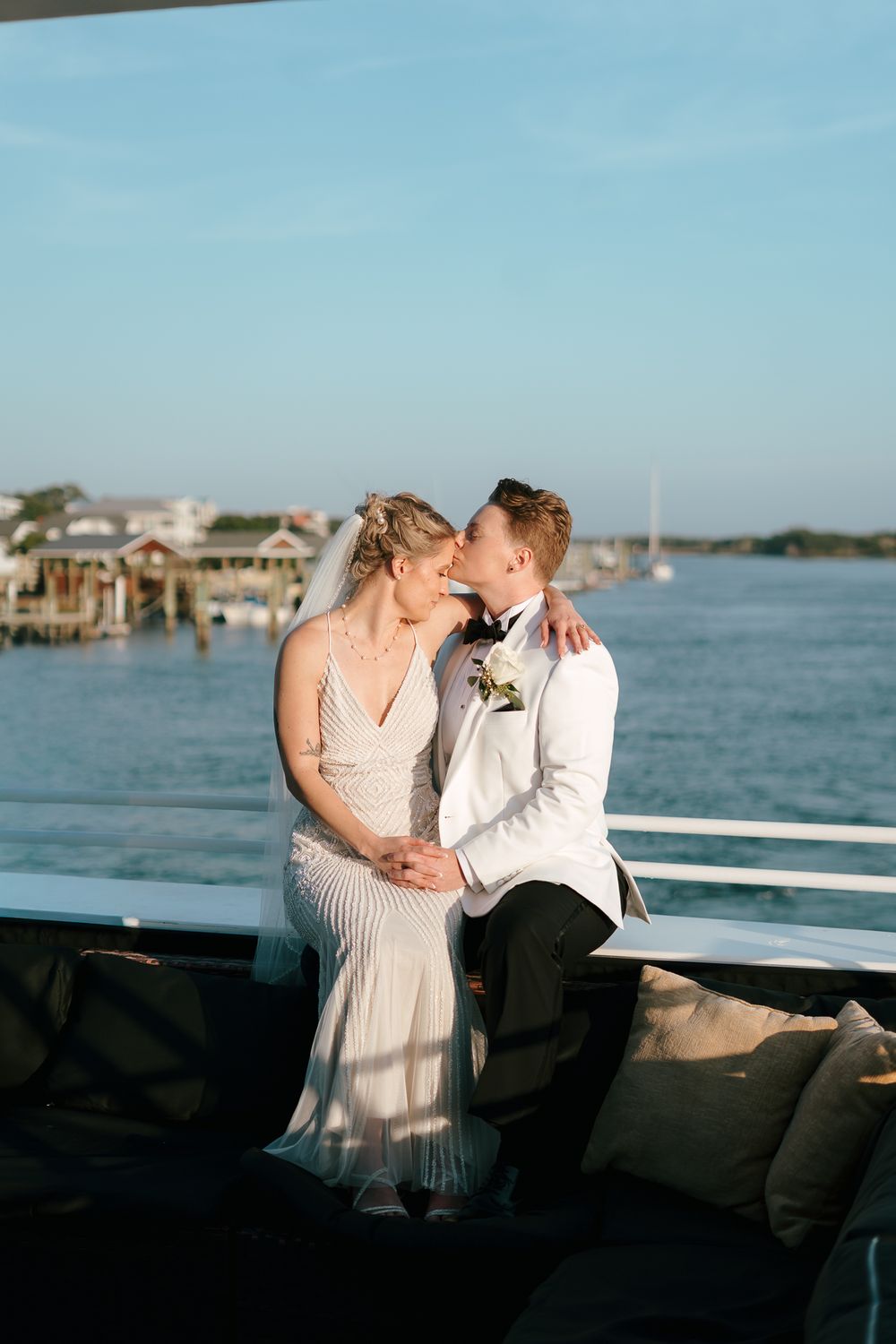 A romantic wedding couple shares a tender moment on a boat deck with waterfront homes and boats in the background.