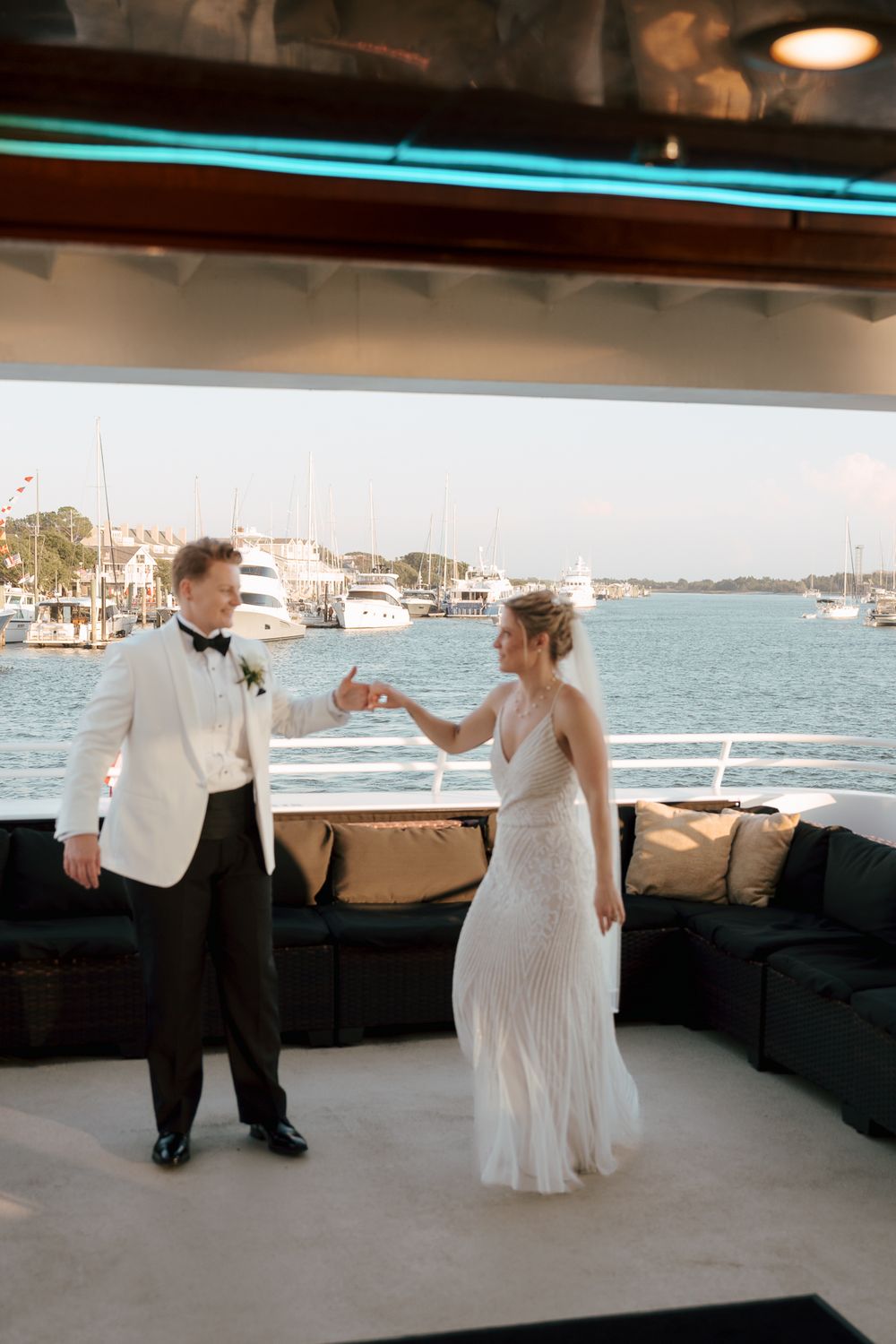 A couple in formal wedding attire dancing together on a yacht deck with a marina and water view in the background.