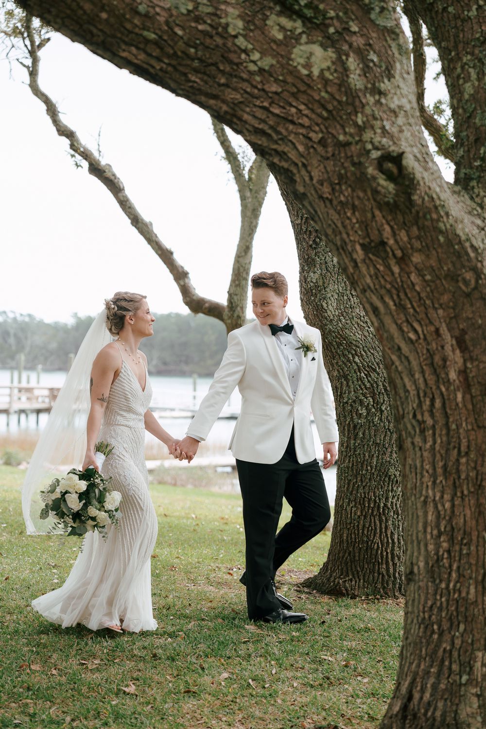 A bride and groom share a romantic moment walking between large oak trees by the water on their wedding day. Beaufort, NC Weddings