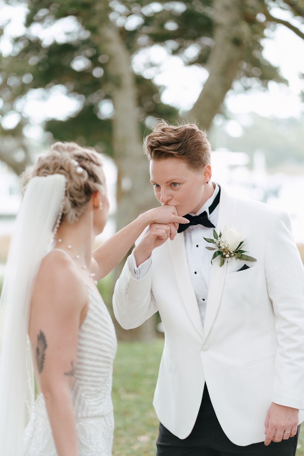 Elegant wedding couple in white attire share intimate moments under tree branches in an outdoor setting. Documentary inspired wedding photo