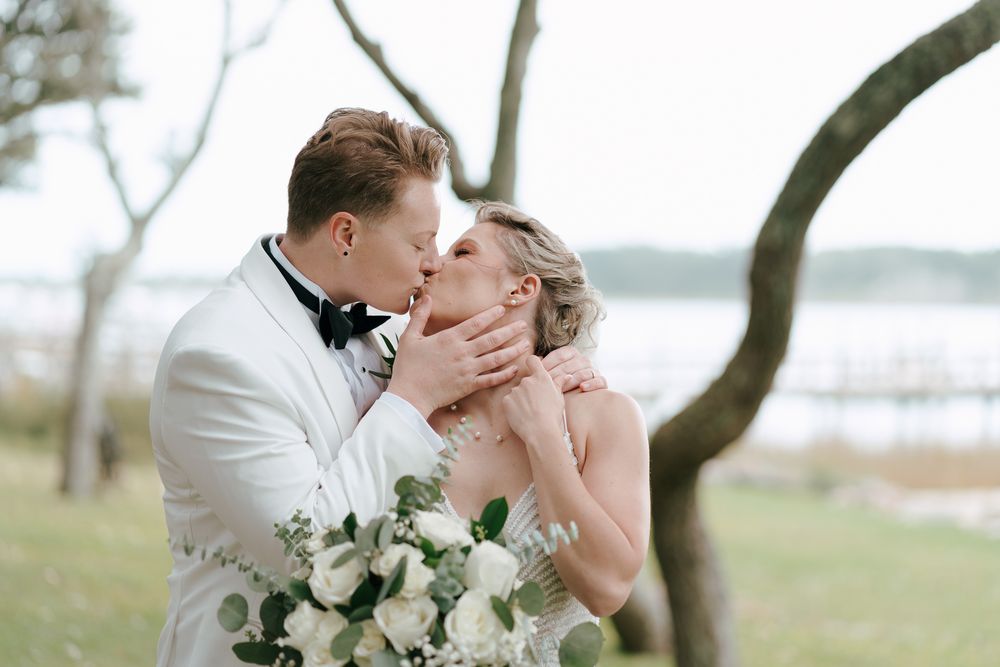A romantic couple embraces during their wedding photoshoot under a tree by the water.