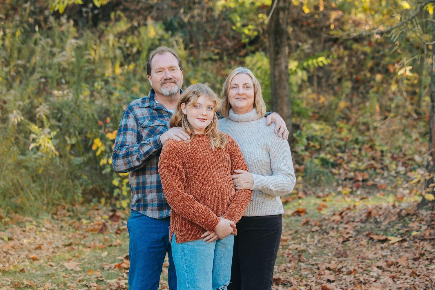A family of three poses together outdoors during autumn with fallen leaves and trees in the background.