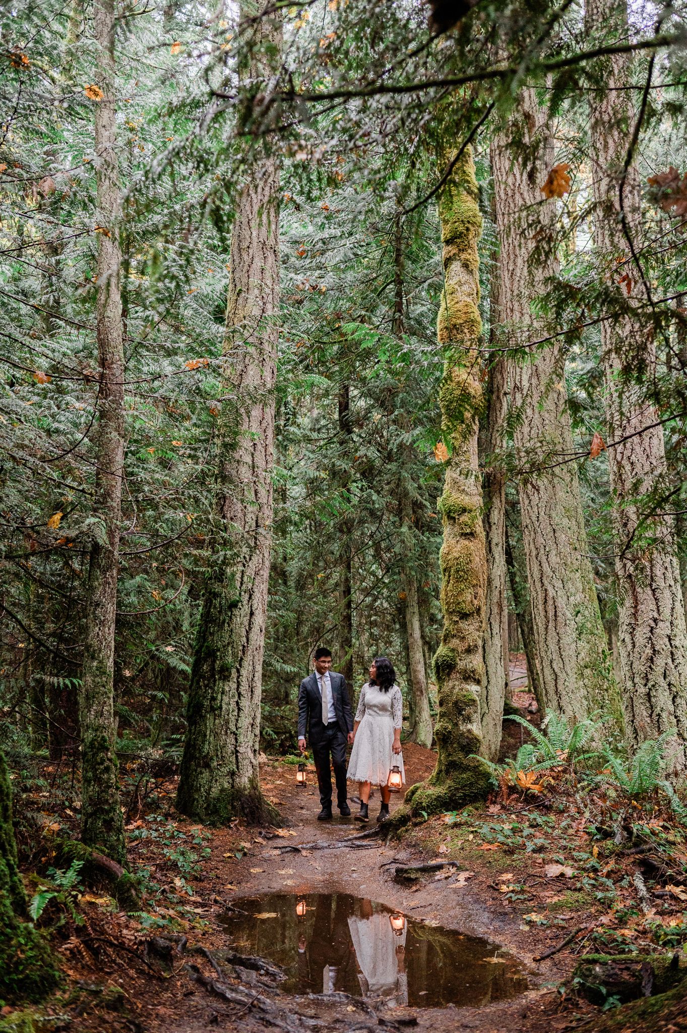 Couple walks along a serene forest path with tall evergreen trees and puddles. Photo by Bainbridge Island Elopement Photographer