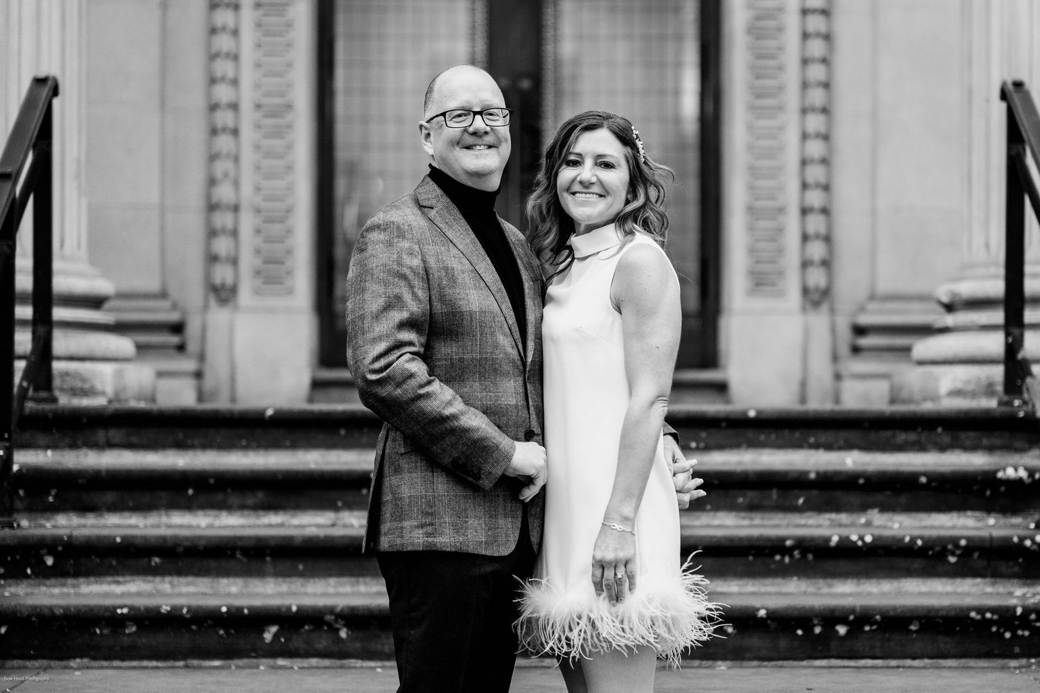 A black and white photo of a couple posing together on a staircase with an urban backdrop.