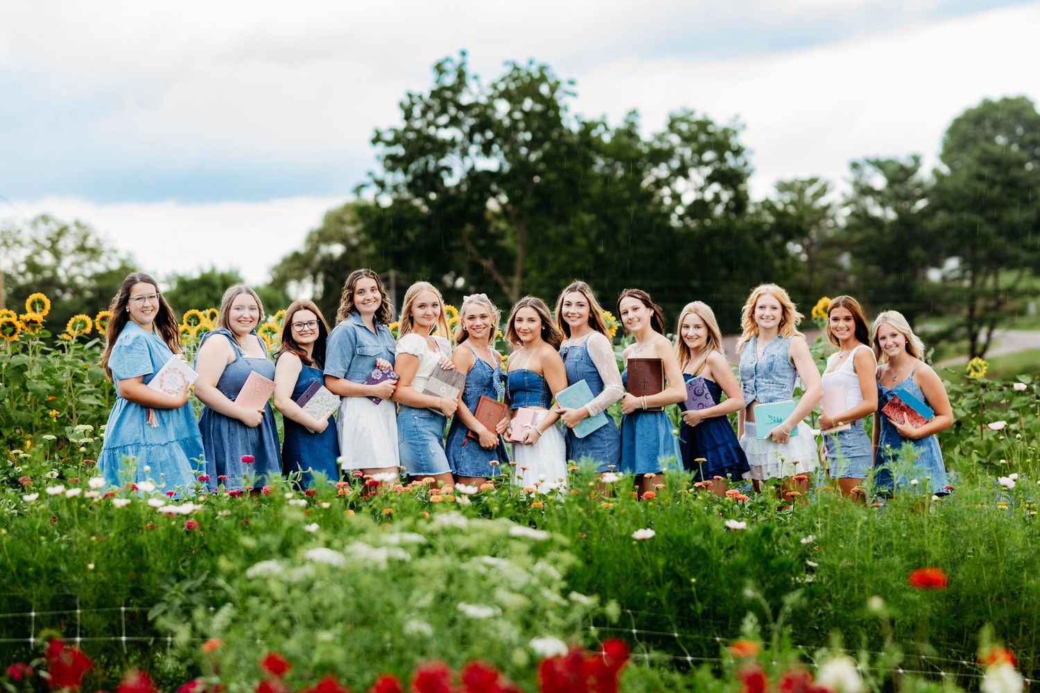 Group of young women in blue dresses holding books while posing in a colorful flower garden.