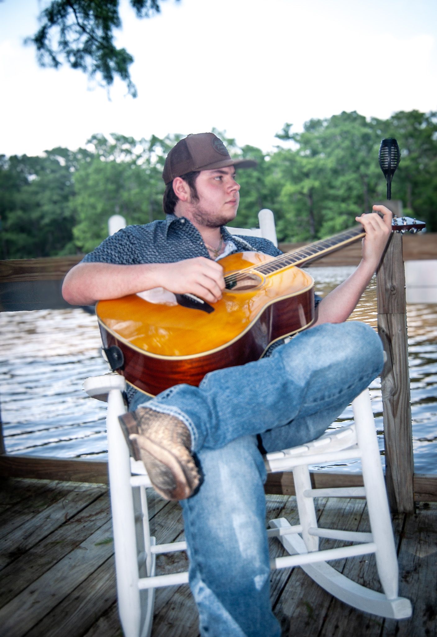 Musician in rocking chair playing acoustic guitar on waterfront dock.