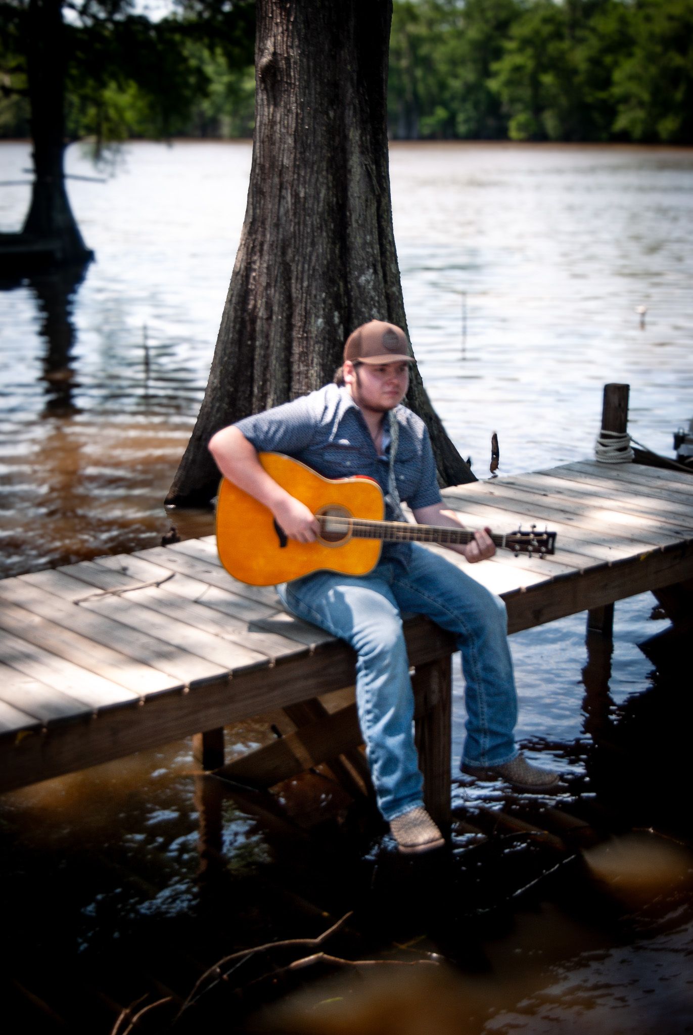 Guitarist sitting on wooden pier with cypress trees and water in background.