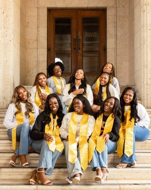 Graduates in yellow stoles pose together on stone steps of academic building.