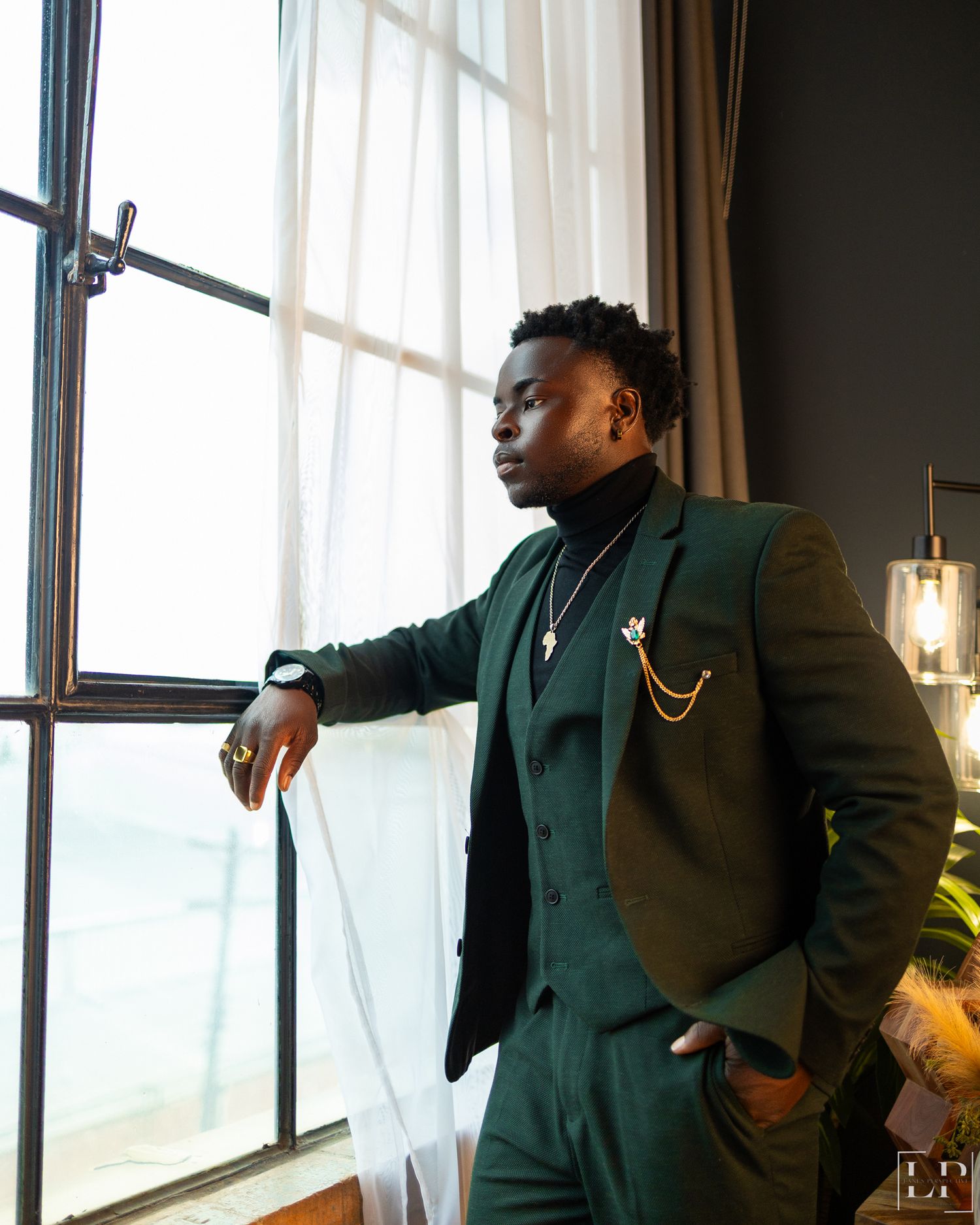 Stylish groomsman in dark suit poses by window in elegant venue.