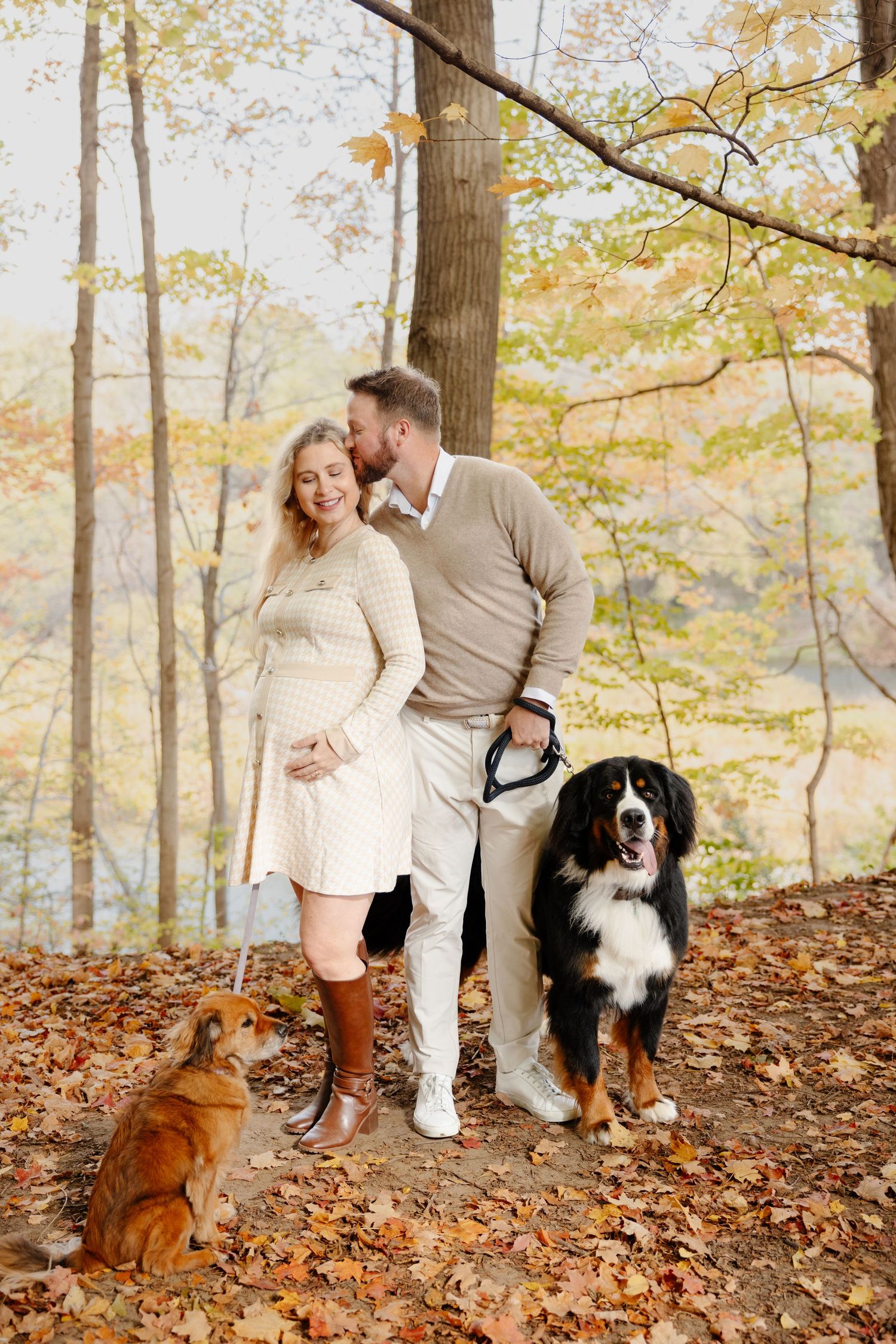Couple poses with their Bernese Mountain Dog and Golden Retriever among fallen autumn leaves in a forest setting.