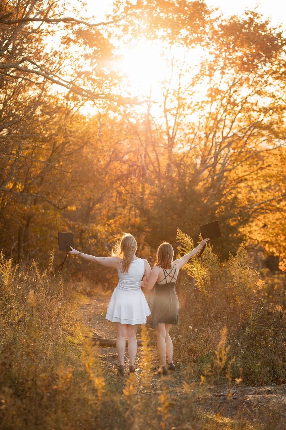 Two figures silhouetted against a golden sunset raise their arms in celebration among autumn trees and tall grass.