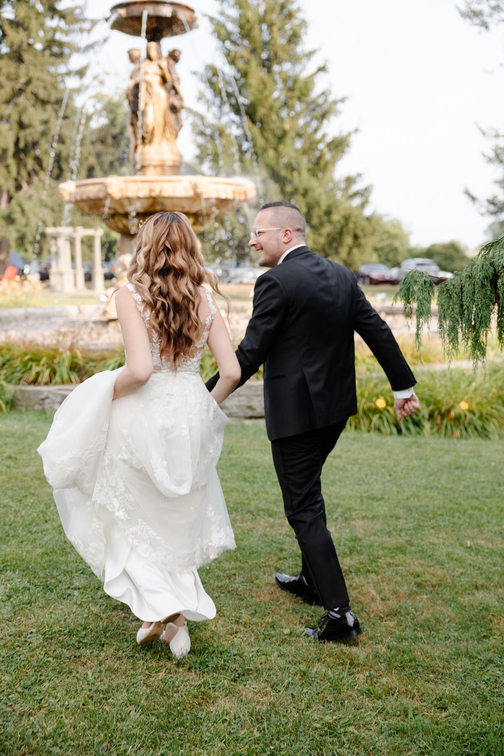 Playful wedding couple running and dancing on a grassy lawn with a fountain in the background.