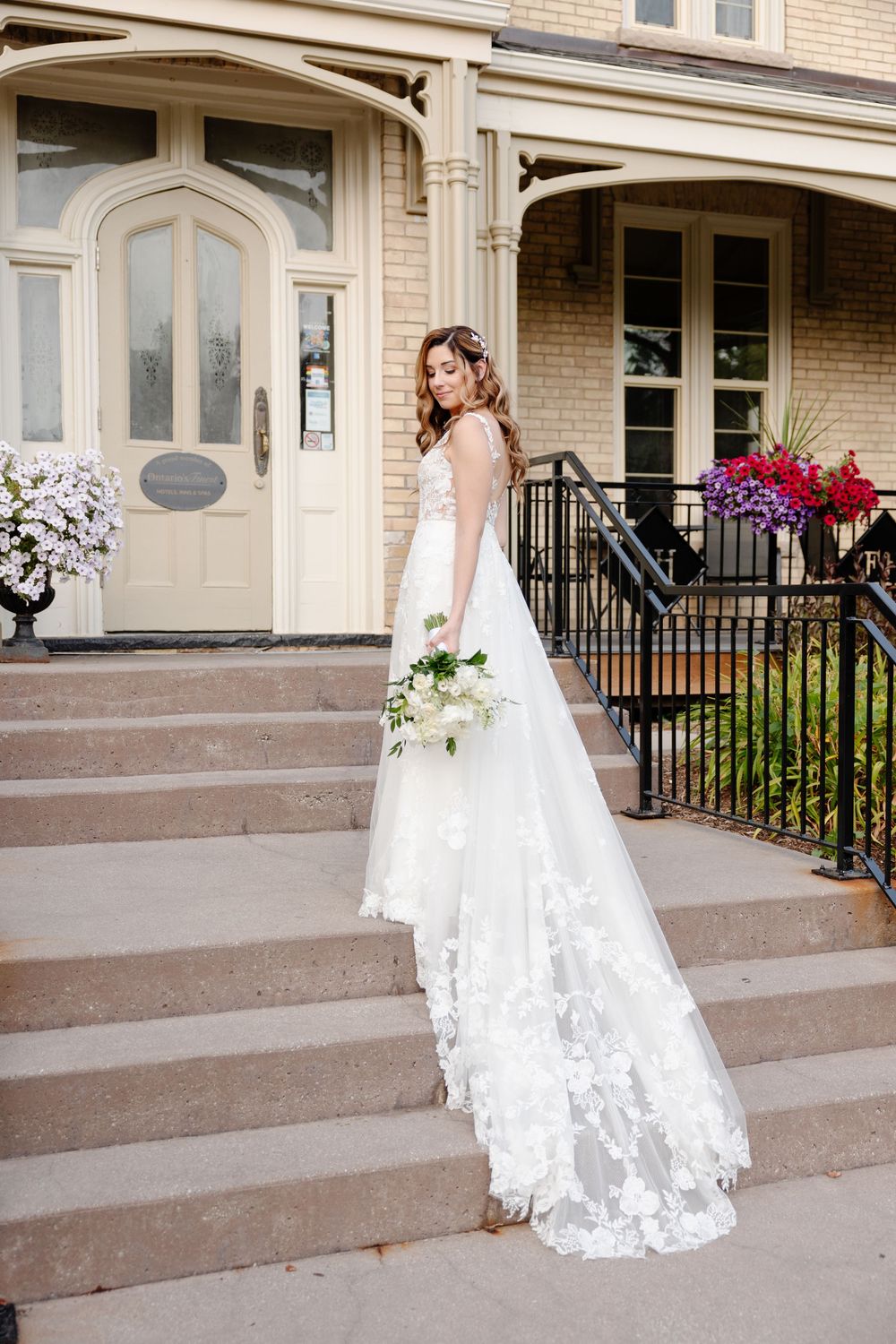 A bride in a flowing white lace wedding gown stands on stone steps outside an elegant brick building with floral decorations.