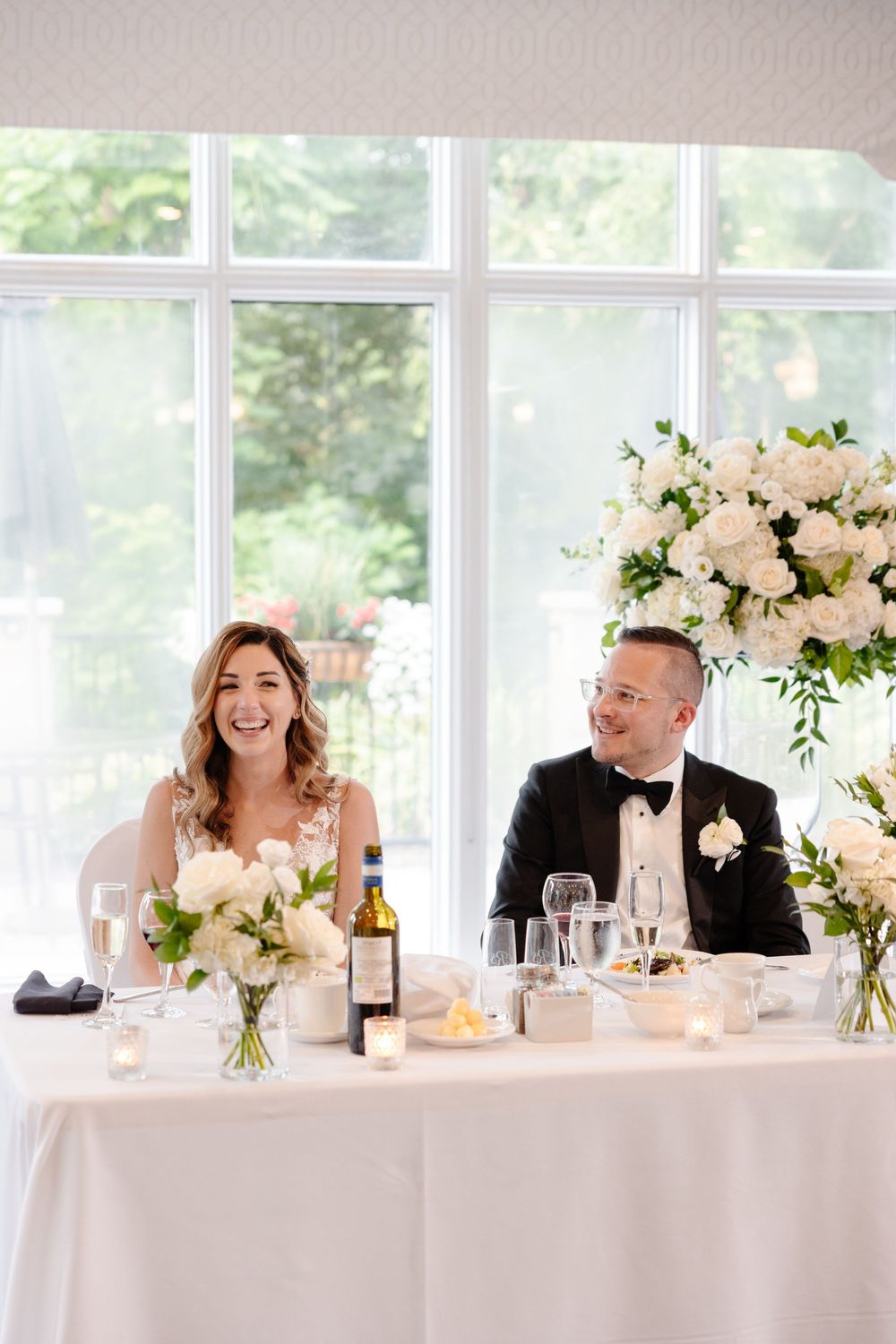 Wedding party members laugh and smile at head table during reception with elegant white floral centerpieces.
