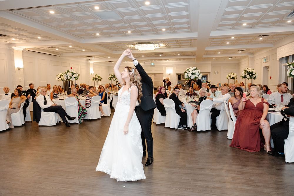 Wedding reception guests dance and celebrate in an elegant white ballroom with crystal chandeliers and floral arrangements.