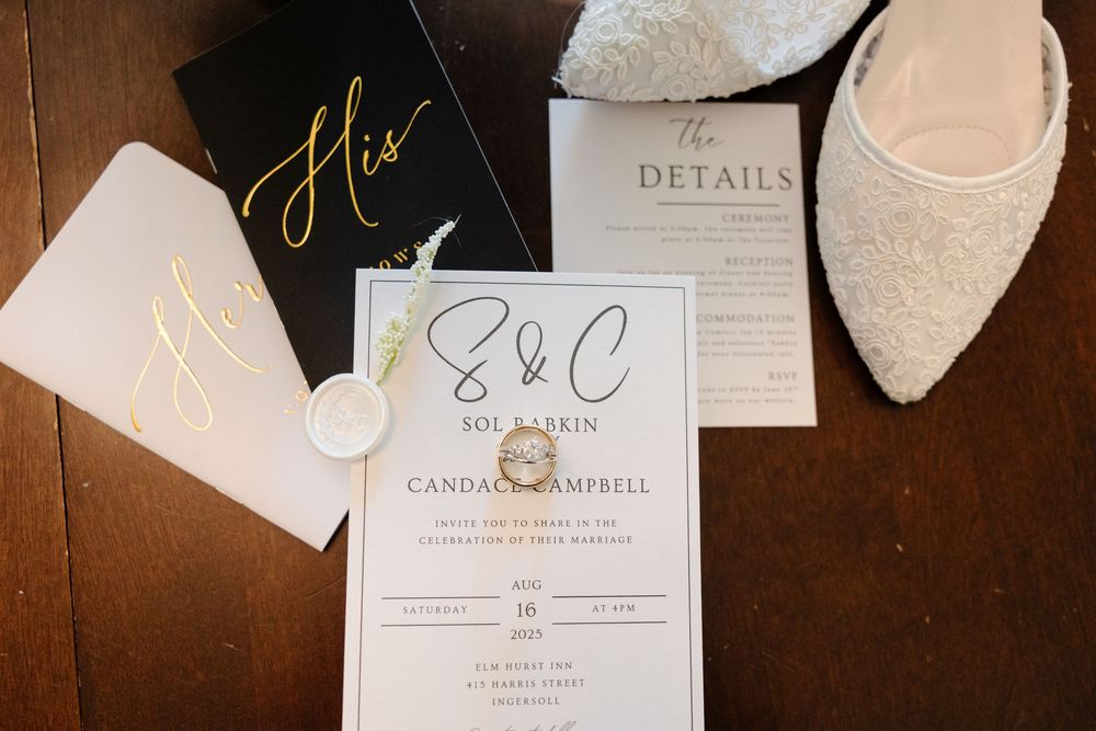 Elegant wedding invitation suite with gold calligraphy lettering and lace bridal shoes on dark wooden surface.