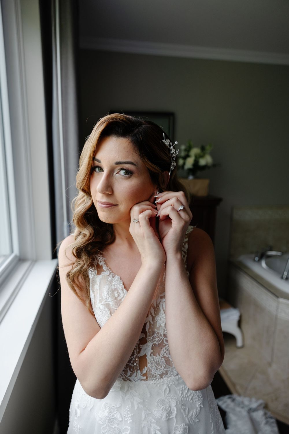 A bride in a white lace wedding dress puts on earrings while standing by a window in natural light.
