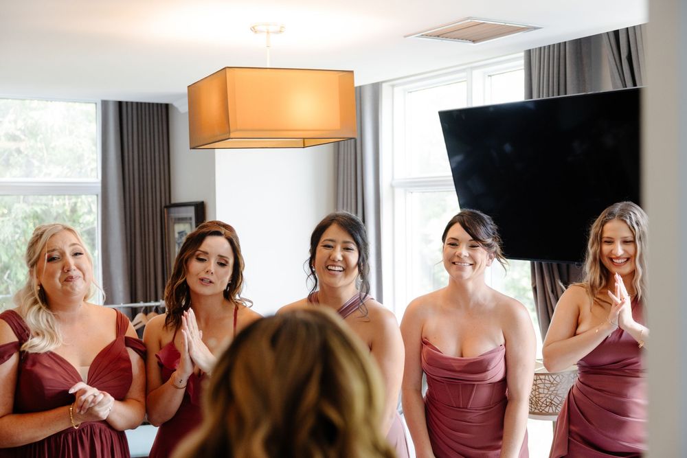 Bridesmaids in burgundy dresses help the bride get ready in a bright hotel room before the wedding ceremony.