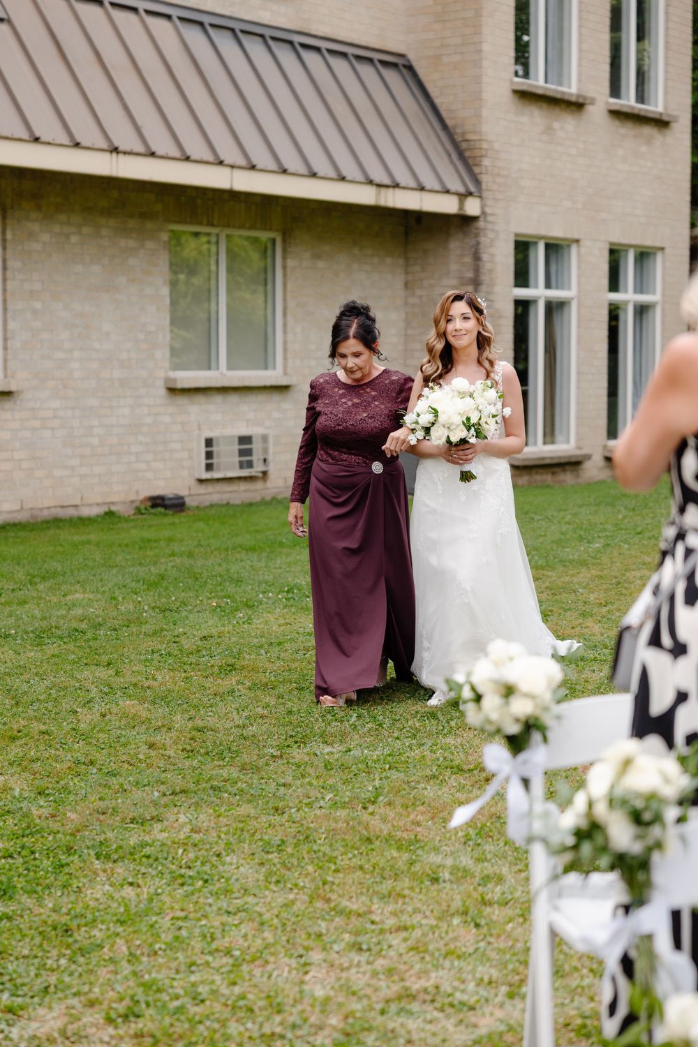 A wedding party stands on a grassy lawn outside a brick building with bridesmaids in burgundy dresses.