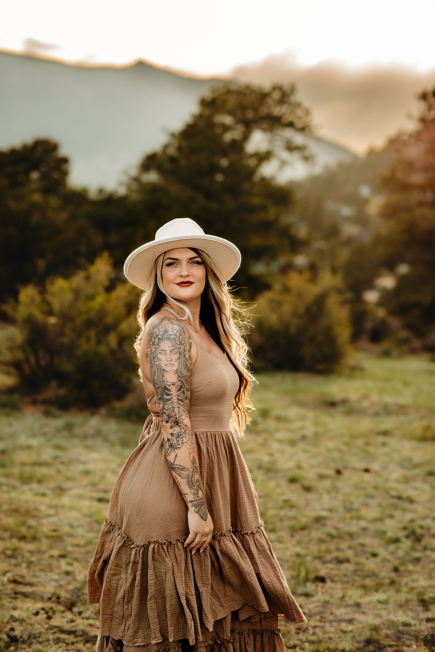 A woman in a brown prairie dress and white hat poses outdoors at sunset with visible tattoo sleeve artwork.