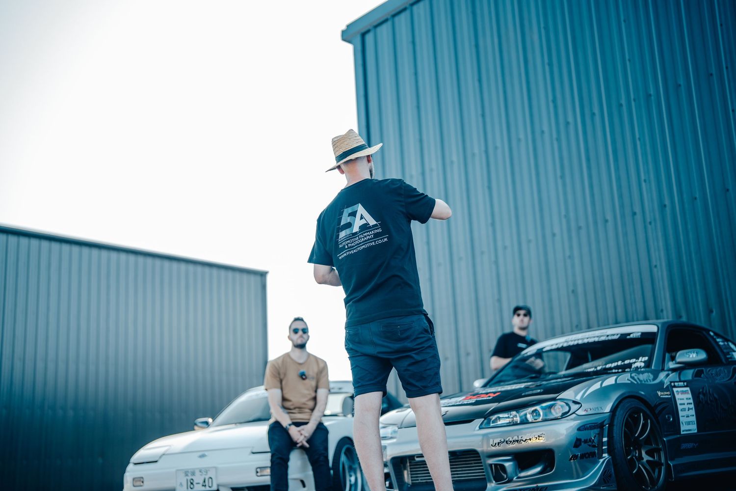 Car enthusiasts gathering outside an industrial building with vehicles on display.