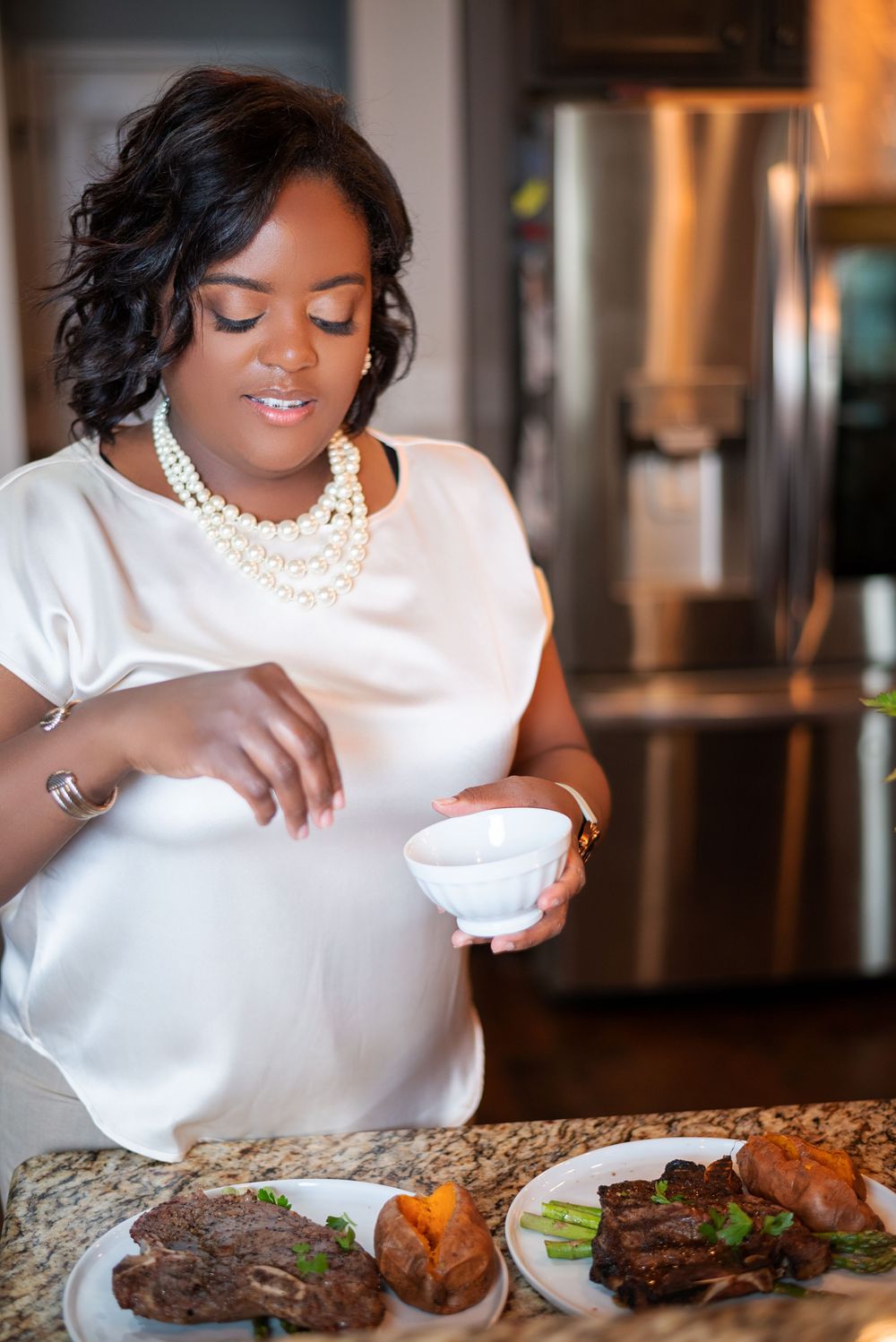 Person in elegant white attire and pearls serving food at a counter with plated steaks and sides.