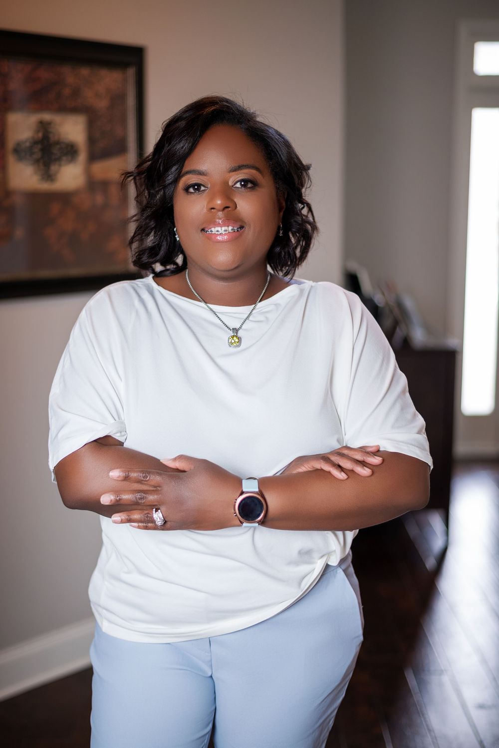 Woman in white top and light blue pants poses confidently in home interior with smart watch and necklace.