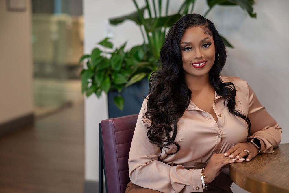 Professional business portrait of someone in a brown blazer sitting in an office setting with indoor plants.