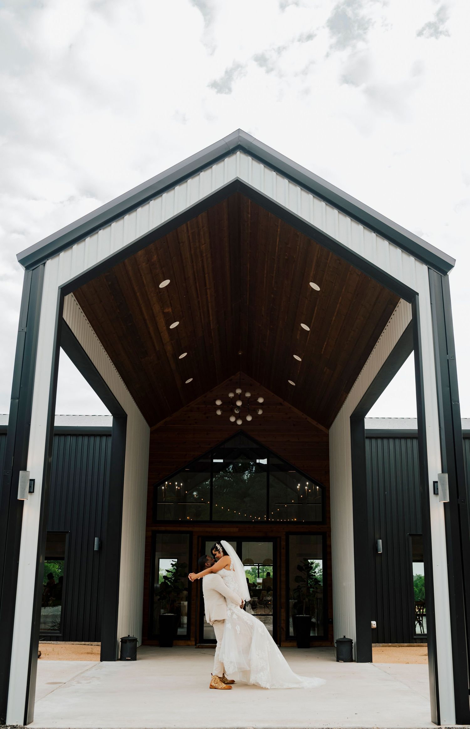 A romantic wedding couple dances together on a gravel path surrounded by lush green trees and sunlight.