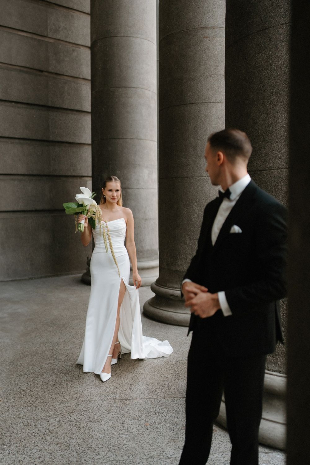 Elegant wedding couple poses between classic stone columns in a formal architectural setting.