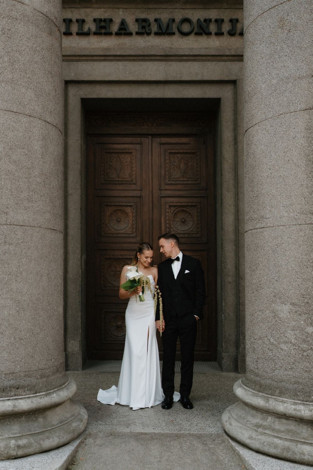 Newlyweds pose intimately between grand stone columns of historic building.