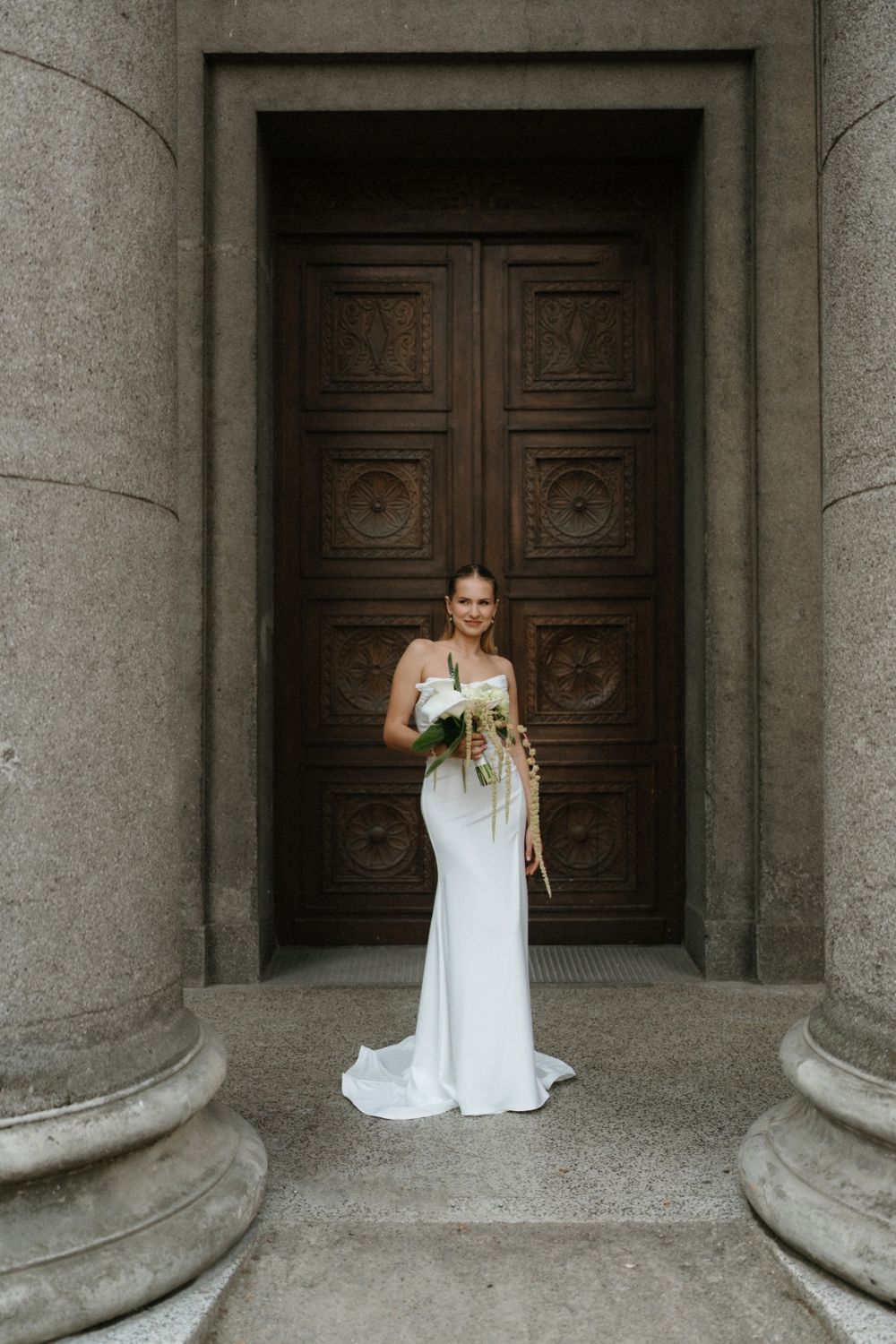 Bride stands elegantly between stone columns holding white floral bouquet.