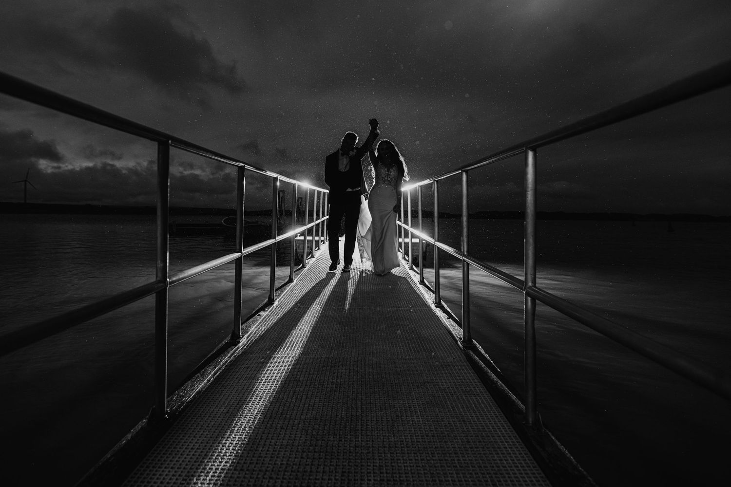A silhouetted figure walks along a dramatic bridge pathway lit by dramatic shadows and light in black and white.
