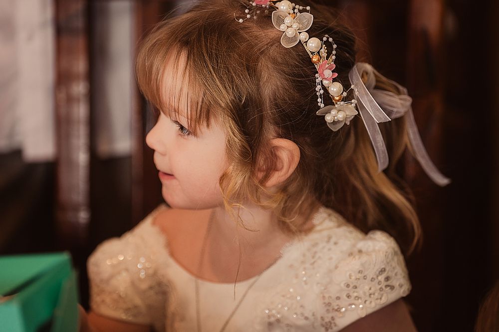 A young child in a white dress with a bow gazes to the side in a warm indoor setting.