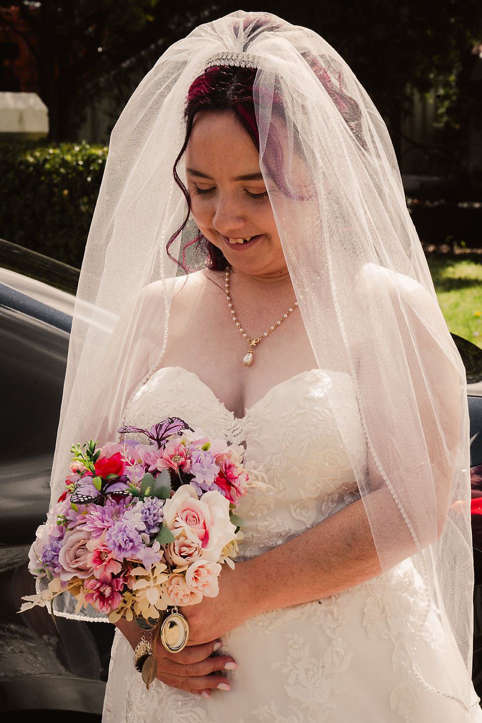 A bride in a white lace wedding dress and veil holds a colorful bouquet of pink and purple flowers in an outdoor setting.