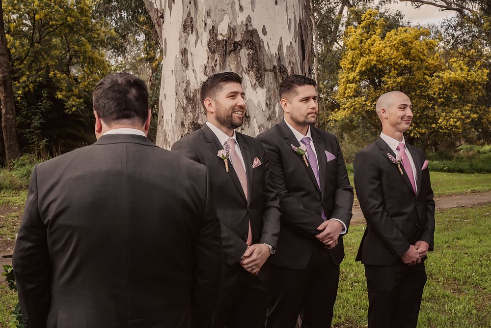 Groomsmen in black suits and pink ties walking in a row through a lush green outdoor setting at a wedding.