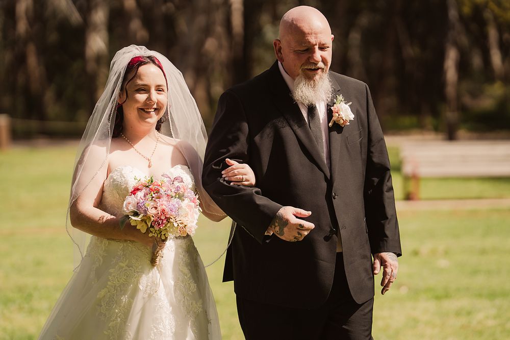 A bride and father walking arm in arm down a path during an outdoor wedding ceremony.