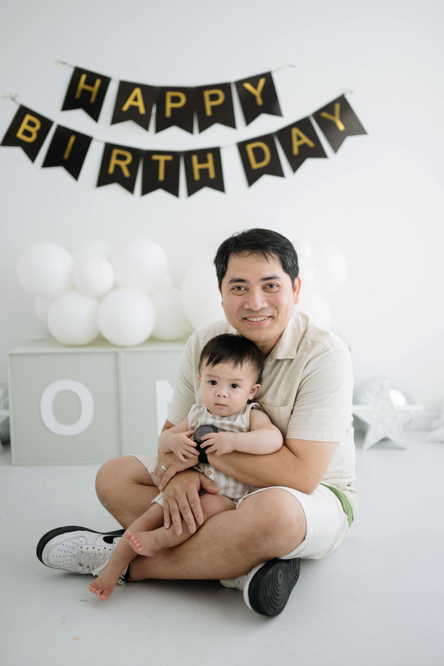 Birthday celebration setup with black and gold banner, white balloons, and decorative blocks in a minimalist white room.